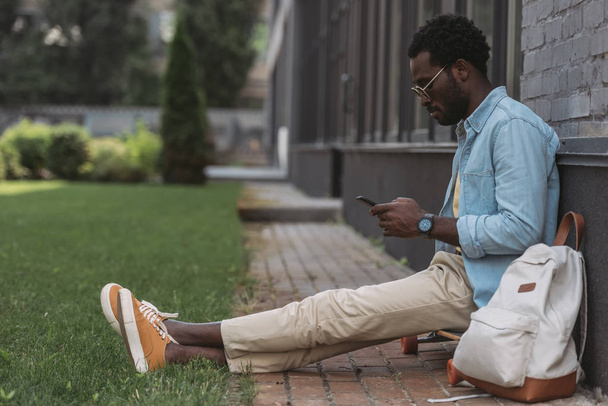 стильний афроамериканський чоловік сидить на дошці і використовує смартфон
 - Фото, зображення