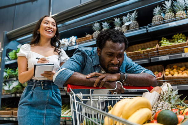 enfoque selectivo de triste afroamericano hombre mirando el carrito de compras cerca de feliz mujer asiática con portátil
 - Foto, imagen