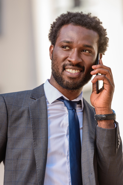 handsome african american businessman talking on smartphone and smiling while looking away - Φωτογραφία, εικόνα