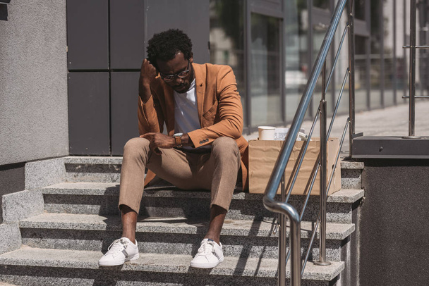 hombre de negocios afroamericano molesto sentado en escaleras cerca de caja de cartón y cogido de la mano cerca de la cabeza
 - Foto, imagen