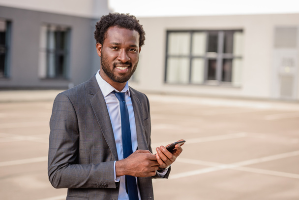 ハンサムなアフリカ系アメリカ人のビジネスマンがスマートフォンを持ち、カメラを見て微笑んでいる - 写真・画像