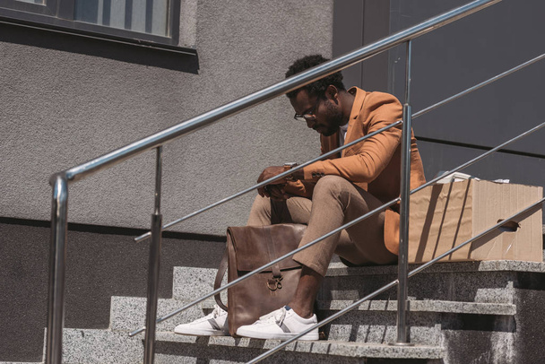 upset, dismissed african american businessman sitting on stairs near cardboard box - Foto, afbeelding