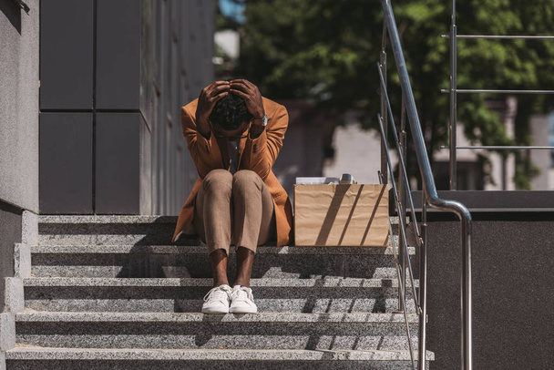 depressief African American zakenman zittend op trappen in de buurt van kartonnen doos en het houden van handen op hoofd - Foto, afbeelding