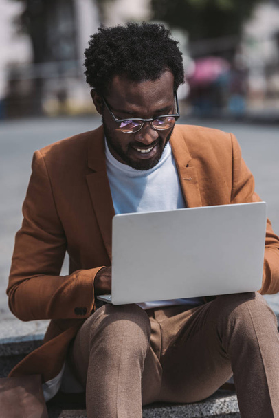 african american businessman in glasses using laptop while sitting on parapet - Foto, Imagen