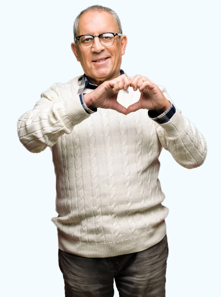 Hombre mayor guapo con gafas y suéter de invierno sonriendo en el amor mostrando el símbolo del corazón y la forma con las manos. Concepto romántico
. - Foto, imagen