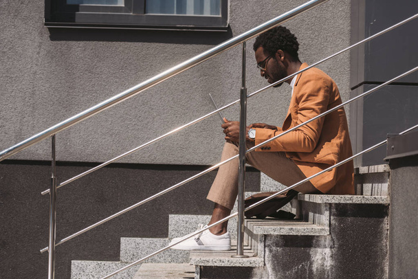вид сбоку на стильного африканского бизнесмена, сидящего на лестнице и использующего ноутбук
 - Фото, изображение