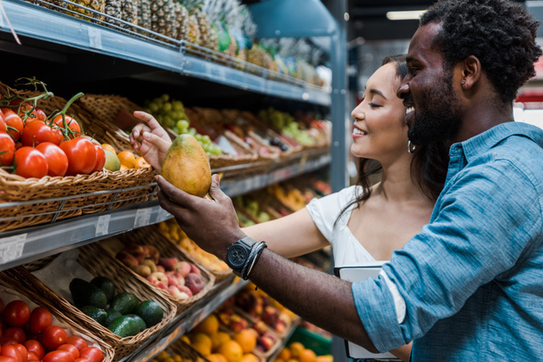 glückliche asiatische Frau schaut auf Früchte in der Nähe fröhlicher afrikanisch-amerikanischer Mann im Geschäft  - Foto, Bild