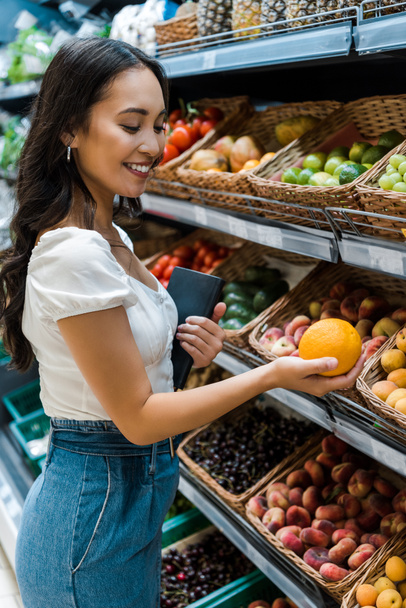 enfoque selectivo de alegre mujer asiática mirando naranja cerca de frutas en la tienda
  - Foto, imagen