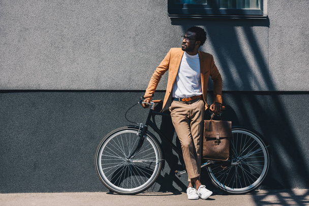 handsome african american businessman standing by wall near bicycle and looking away - Fotoğraf, Görsel