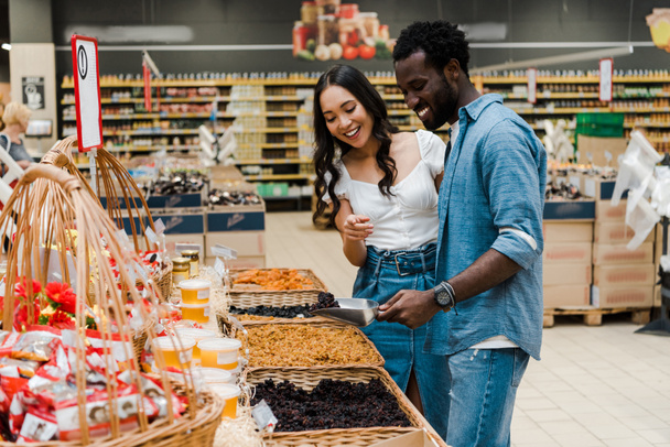 felice uomo africano americano in possesso di scoop metallo con uva passa vicino donna asiatica che punta con il dito in negozio
  - Foto, immagini