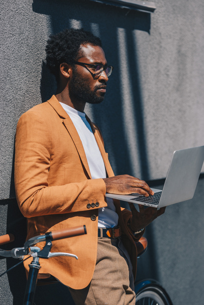 thoughtful african american businessman in glasses using laptop while standing with bicycle near wall - Fotoğraf, Görsel
