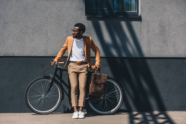 自転車の近くの壁に立ちながら目をそらすハンサムなアフリカ系アメリカ人ビジネスマン - 写真・画像
