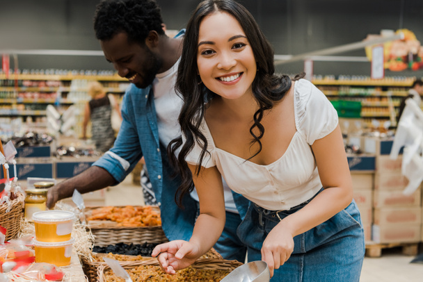 feliz asiático mujer mirando a cámara cerca alegre africano americano hombre mirando miel tarros en supermercado
  - Foto, Imagen
