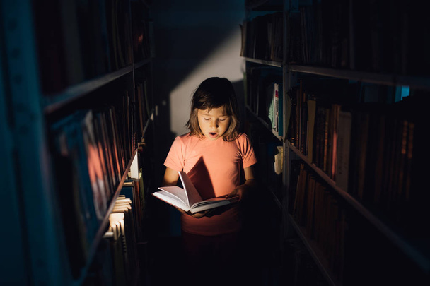 klein meisje leest het boek, is bang, heeft open mond. houdt een boek in zijn hand, leert in de bibliotheek, binnenshuis, 1 - Foto, afbeelding