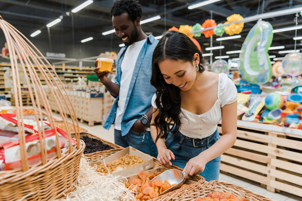 selective focus of happy asian woman looking at dried apricots near cheerful african american man holding honey jar in supermarket  - Photo, Image