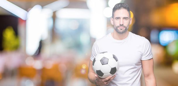 Νέος άνθρωπος που κρατά την μπάλα ποδοσφαίρου ποδοσφαίρου πέρα από το απομονωμένο υπόβαθρο με αυτοπεποίθηση έκφραση στο έξυπνο πρόσωπο σκέφτεται σοβαρά - Φωτογραφία, εικόνα