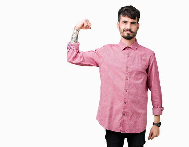 Fiatal jóképű ember visel rózsaszín inget át elszigetelt háttér erős személy mutató kar izom, magabiztos és büszke erő - Fotó, kép