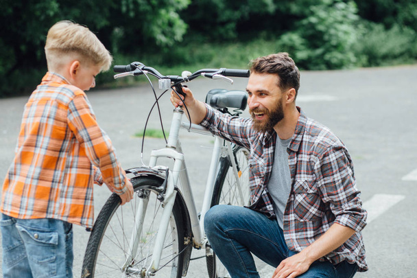 Όμορφος μπαμπάς και το χαριτωμένο μικρό γιο του με ποδήλατο στο πάρκο, κοιτάζοντας ο ένας τον άλλο και χαμογελώντας. Ο πατέρας εξετάζει το ποδήλατο του γιου του.. - Φωτογραφία, εικόνα