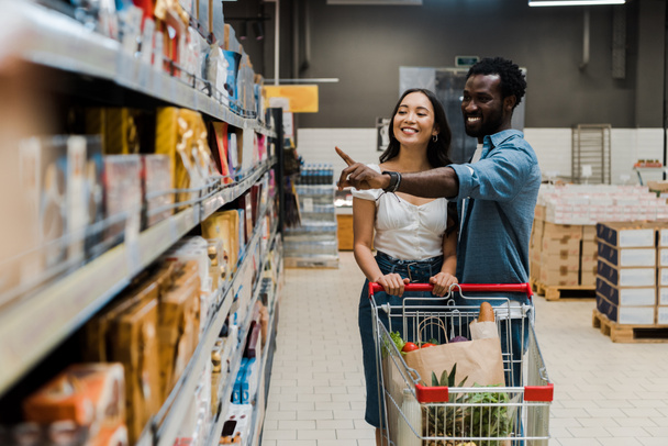 foco seletivo de mulher asiática feliz perto de homem americano africano alegre apontando com o dedo para prateleiras no supermercado
  - Foto, Imagem