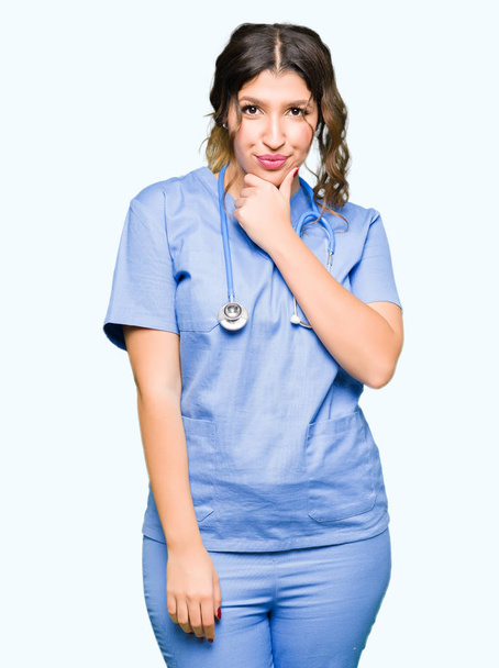 Jovem mulher médica adulta vestindo uniforme médico olhando confiante para a câmera com sorriso com braços cruzados e mão levantada no queixo. Pensar positivo
. - Foto, Imagem