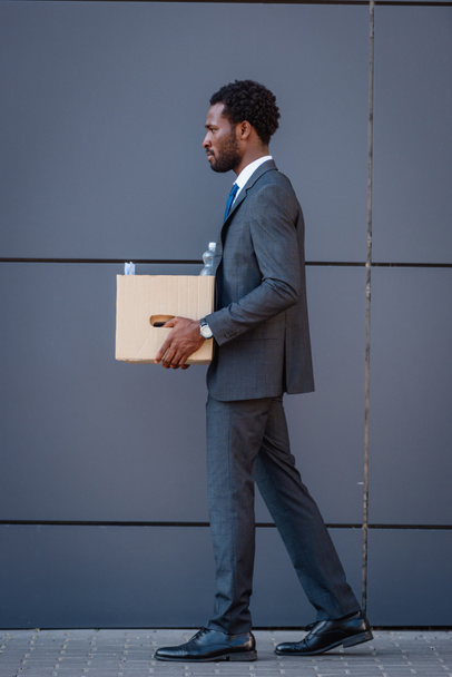 side view of fired african american businessman holding carton box while walking on street - Φωτογραφία, εικόνα