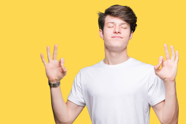 Jonge knappe man dragen casual wit t-shirt over geïsoleerde achtergrond ontspannen en lachend met ogen gesloten doen meditatie gebaar met vingers. Yoga concept. - Foto, afbeelding