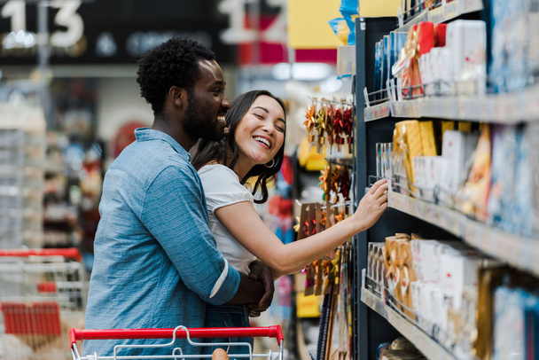 selective focus of cheerful african american man standing with asian woman smiling near groceries in supermarket  - Фото, изображение