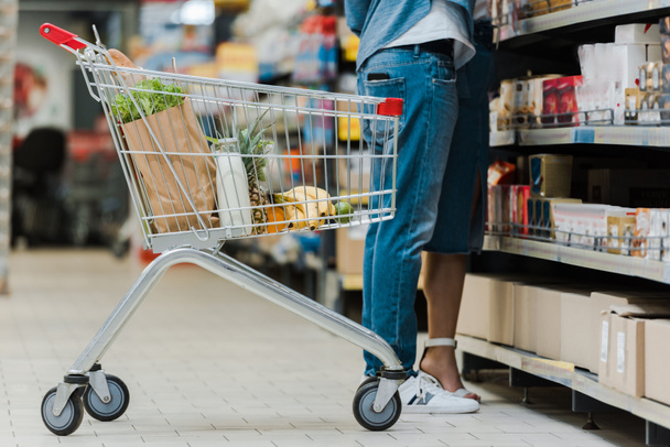cropped view of man standing with woman near shopping cart with groceries in supermarket  - Photo, Image