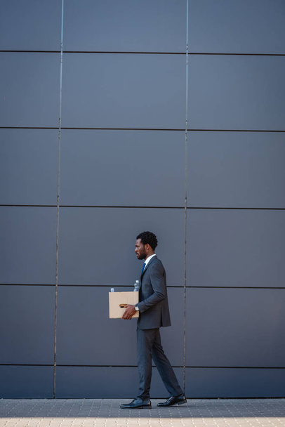 Seitenansicht eines gefeuerten afrikanisch-amerikanischen Geschäftsmannes, der einen Karton bei sich trägt, während er an der Wand entlang läuft - Foto, Bild