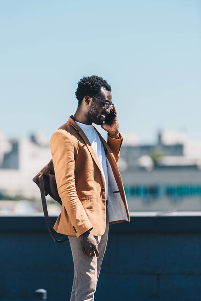κομψός Αφρικανός Αμερικανός επιχειρηματίας μιλάει στο smartphone και χαμογελά στην ταράτσα - Φωτογραφία, εικόνα