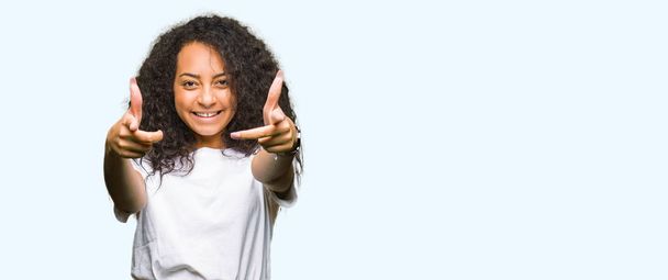 Молодая красивая девушка с вьющимися волосами в повседневной белой футболке указывает на тебя и камеру пальцами, улыбаясь позитивно и весело
 - Фото, изображение