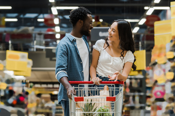 szczęśliwy African amerykański mężczyzna patrząc w Asian Dziewczyna wskazując blisko koszyk w supermarket  - Zdjęcie, obraz