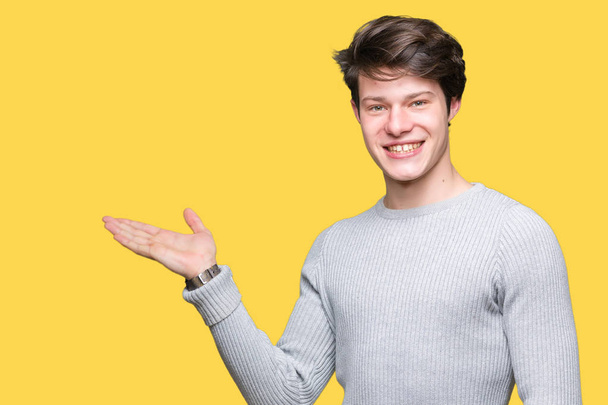 Young handsome man wearing winter sweater over isolated background smiling cheerful presenting and pointing with palm of hand looking at the camera. - Photo, Image