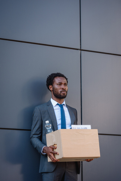 dismissed, upset african american businessman holding carton box and looking away while standing near wall  - Fotoğraf, Görsel