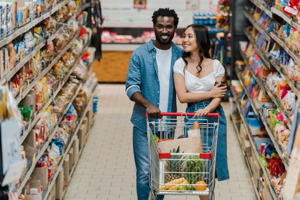 スーパーマーケットでショッピングカートを持って歩く幸せなアジアの女性とアフリカ系アメリカ人男性の選択的な焦点  - 写真・画像