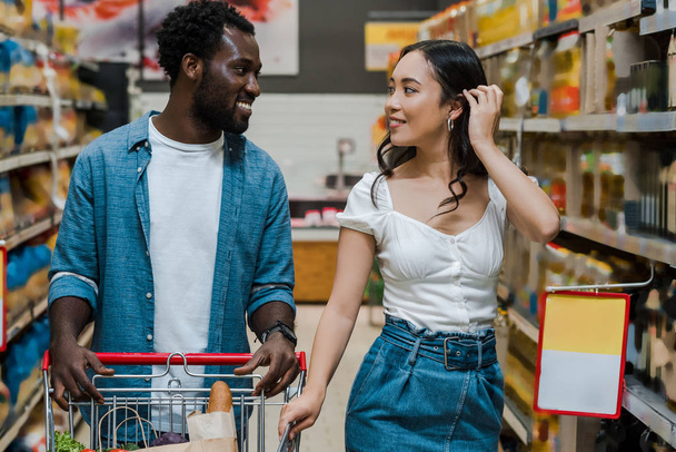 felice giovane donna asiatica toccare i capelli e guardando l'uomo africano americano in negozio
  - Foto, immagini