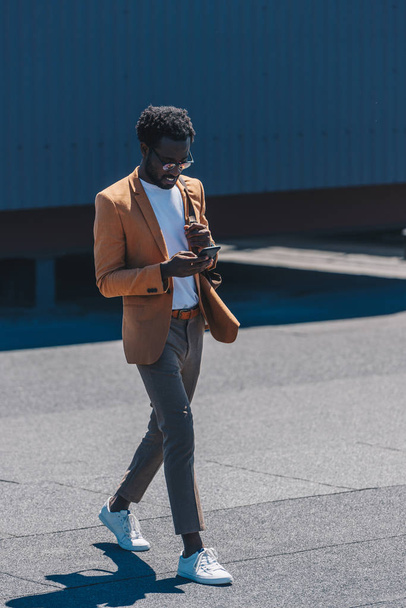 sonriente y elegante hombre de negocios afroamericano usando un teléfono inteligente mientras camina en la azotea
 - Foto, Imagen
