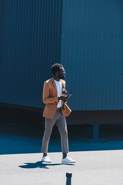 スタイリッシュなアフリカ系アメリカ人ビジネスマンがスマートフォンを持ち、屋上を歩きながら目をそらす - 写真・画像