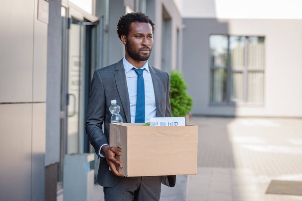 fired, upset african american businessman looking away while holding cardboard box - Фото, изображение