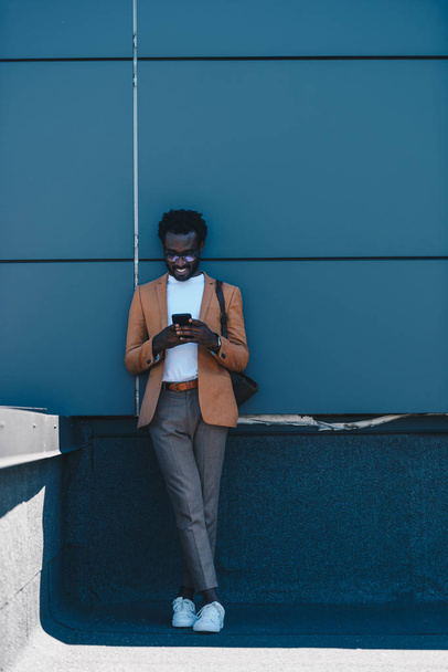 handsome, smiling african american businessman standing on rooftop and using smartphone - Foto, Imagem
