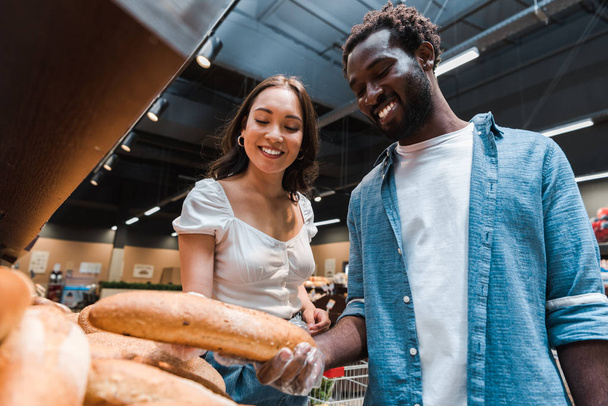 Low-Winkelaufnahme eines afrikanisch-amerikanischen Mannes mit Baguette im Supermarkt in der Nähe einer asiatischen Frau  - Foto, Bild