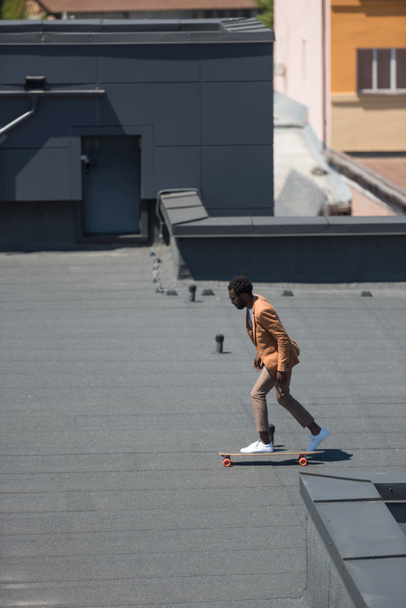 stylischer afrikanisch-amerikanischer Geschäftsmann auf Longboard auf sonnigem Dach - Foto, Bild
