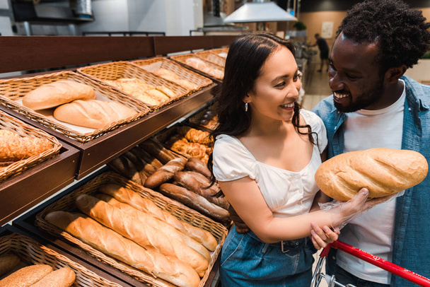 glücklich asiatische Frau hält Brot in der Nähe fröhlichen afrikanisch-amerikanischen Mann  - Foto, Bild