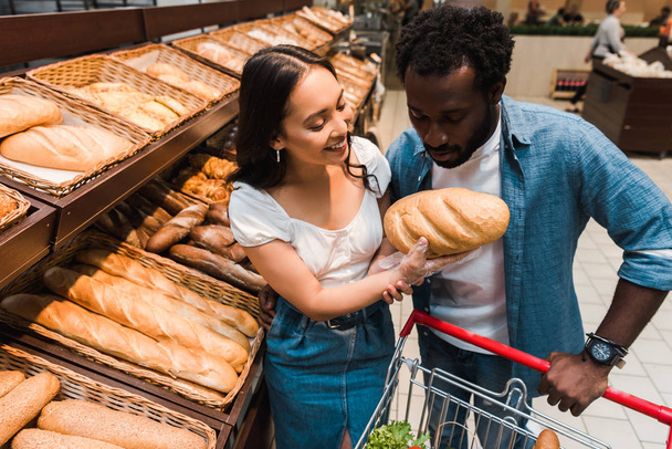 african american man looking at bread in hand of cheerful asian woman  - Photo, Image
