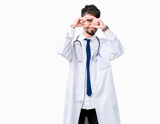 Young doctor man wearing hospital coat over isolated background Doing heart shape with hand and fingers smiling looking through sign - Valokuva, kuva