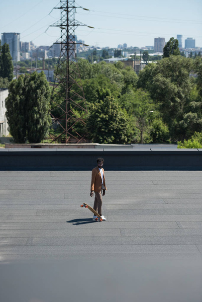 stylish african american businessman with longboard on rooftop with green trees on background - Φωτογραφία, εικόνα