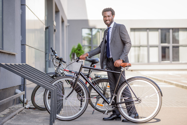 handsome african american businessman smiling at camera while taking bike from parking  - Foto, Imagem