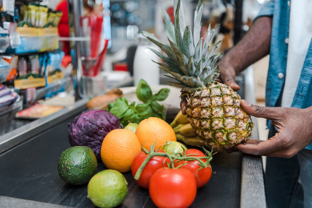 cropped view of african american man holding pineapple near supermarket counter with groceries - 写真・画像