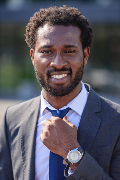 handsome, cheerful african american businessman touching tie and smiling at camera - 写真・画像