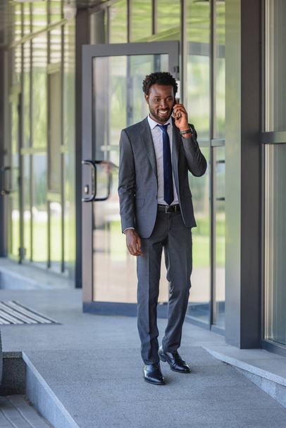 smiling african american businessman talking on smartphone while walking along office buiding - Photo, Image